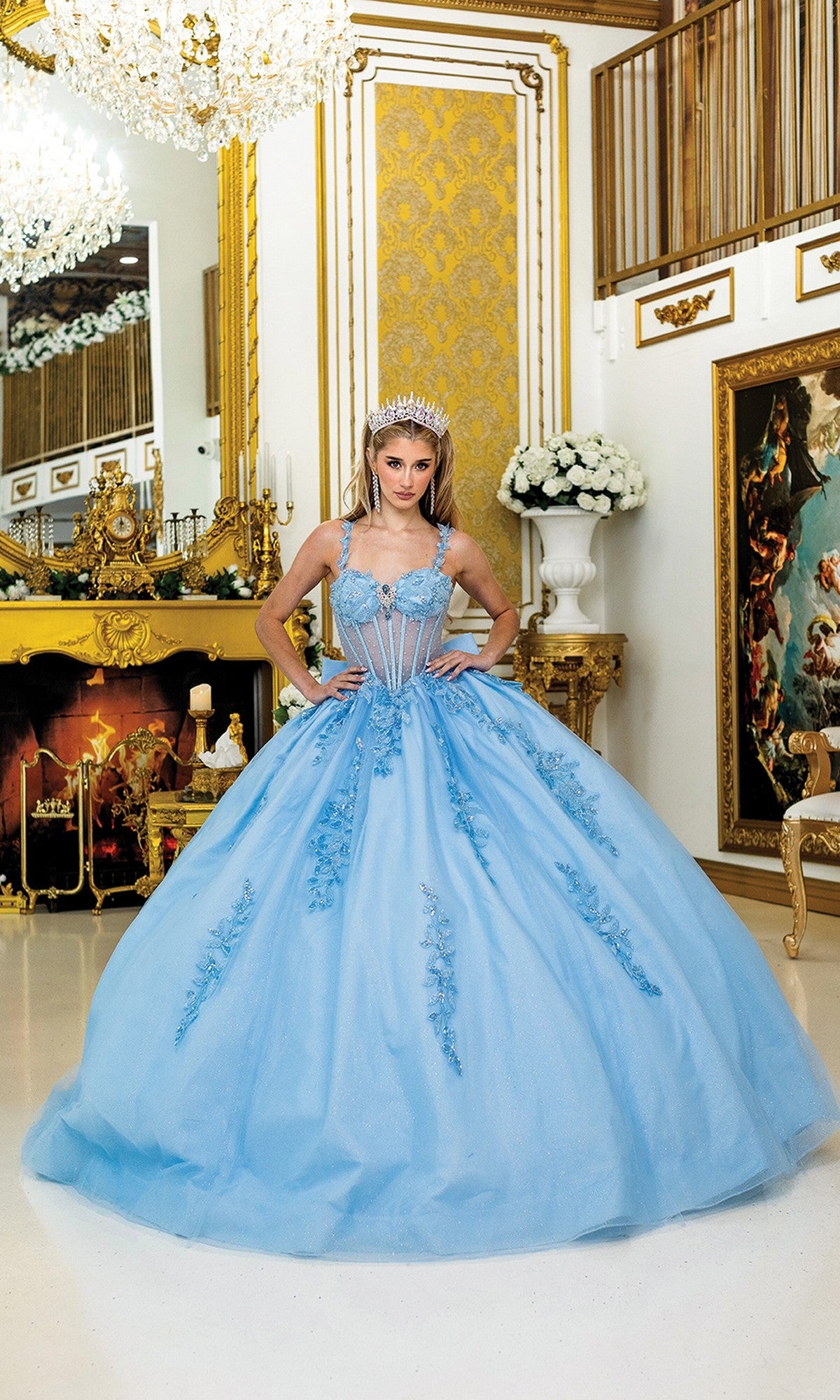 A person in the Quinceanera Dress 5046 By Dancing Queen, featuring a blue ball gown and corset bodice with tiara, stands in an ornate gold-accented room with a fireplace, chandeliers, and classical decor.
